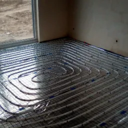 Układanie rur ogrzewania podłogowego na folii z nadrukiem siatki, widok z góry, surowe ściany w tle, widoczne okno z widokiem na zewnątrz.