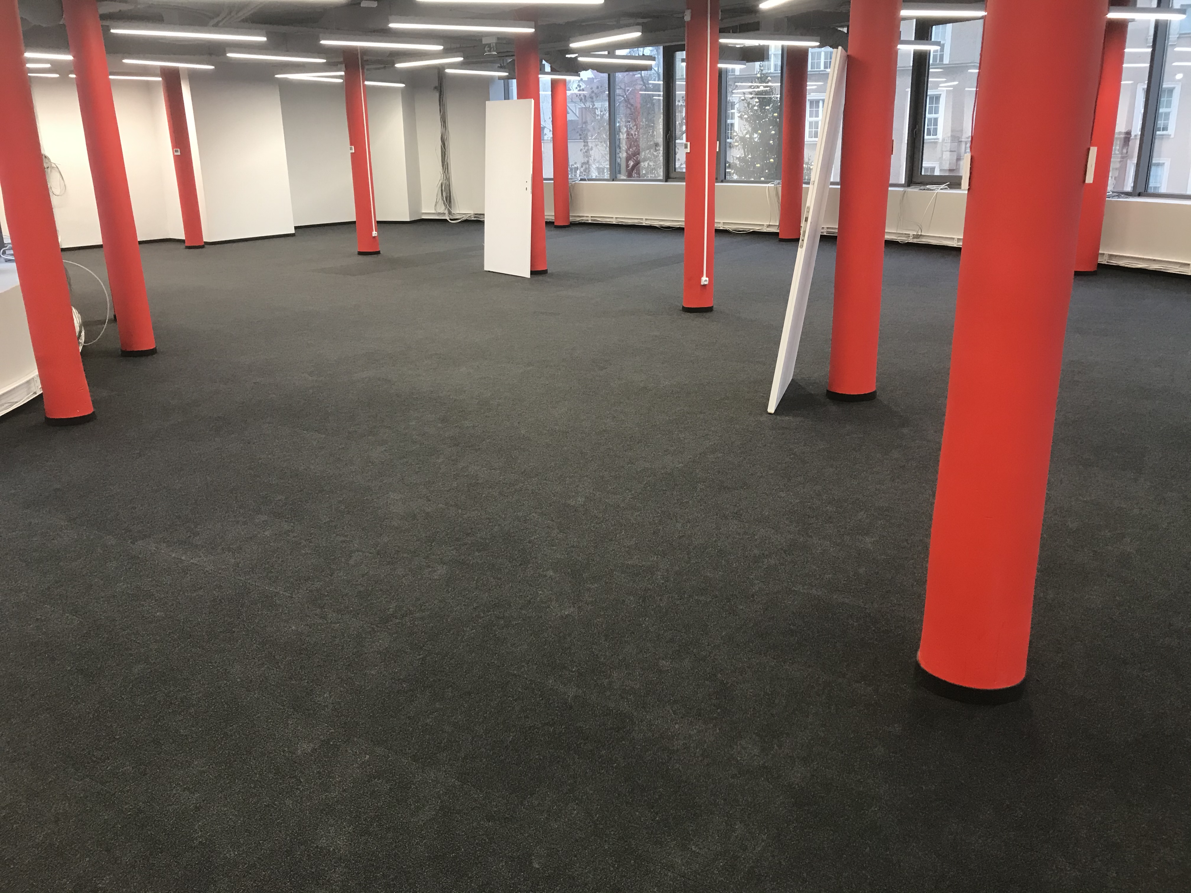Wnętrze biurowe z ciemną wykładziną dywanową, czerwonymi kolumnami i widokiem na budynki za oknem.