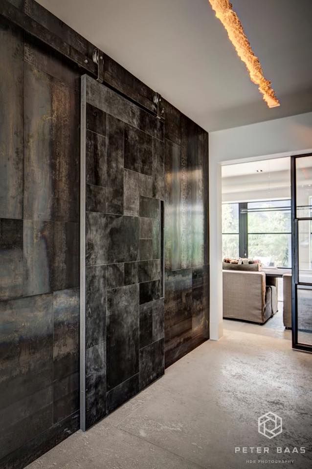 Wnętrze z przesuwnymi drzwiami w stylu industrialnym, wykonanymi z metalowych paneli o zróżnicowanej fakturze i kolorystyce, zawieszonymi na widocznych szynach; w tle widoczny salon z jasną sofą...