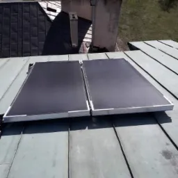 Dwa panele słoneczne zamontowane na metalowym dachu, widok z góry, w tle komin i trawnik.