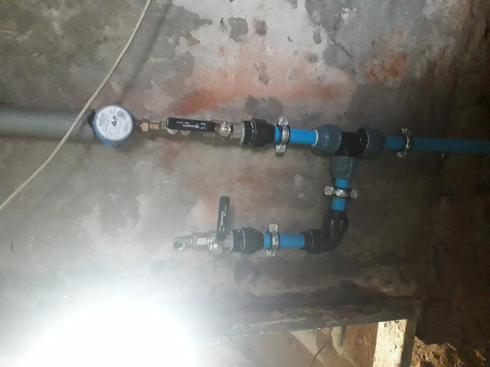 Fragment instalacji wodnej z licznikiem i zaworami, rury niebieskie i czarne, mocowane metalowymi klamrami, na tle surowej ściany z widocznymi śladami wilgoci.