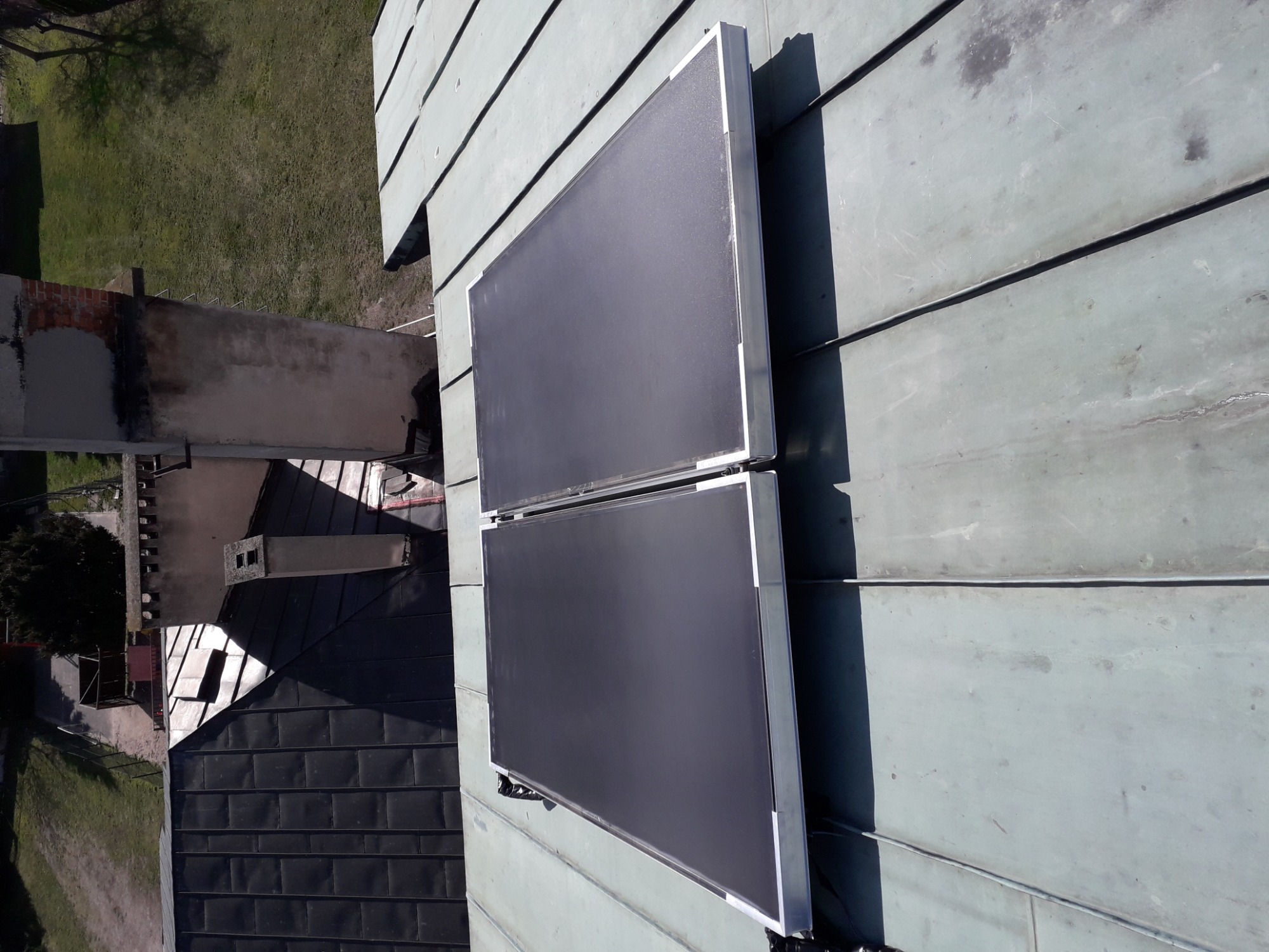 Dwa panele słoneczne na metalowym dachu, widok z góry, w tle komin i zieleń.