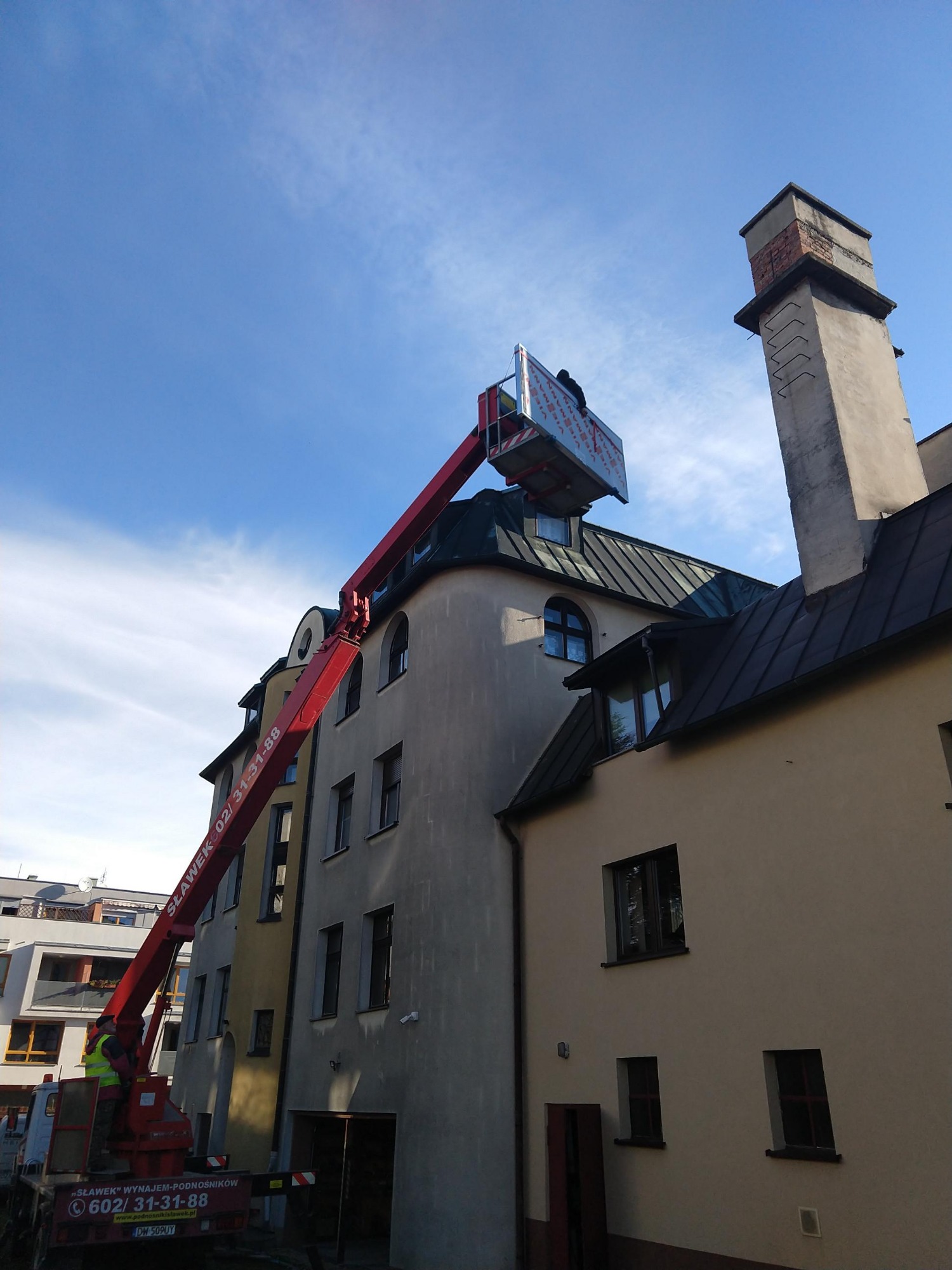 Pracownik na podnośniku koszowym przy wysokim kominie na dachu kamienicy, widok z dołu na tle błękitnego nieba.