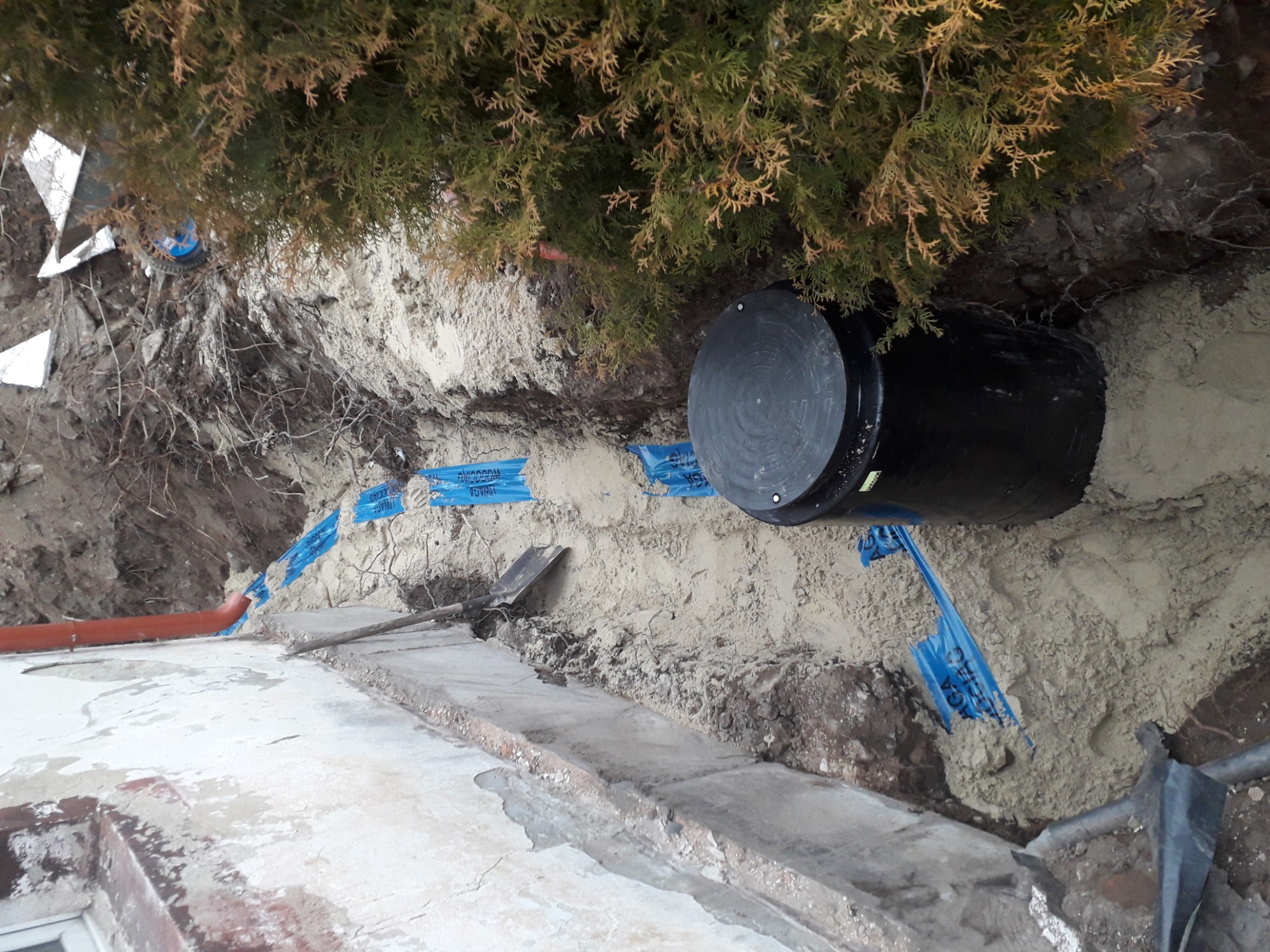 Czarny zbiornik retencyjny częściowo zasypany piaskiem w wykopie, widoczne niebieskie taśmy zabezpieczające, rura kanalizacyjna i fragment fundamentu budynku.