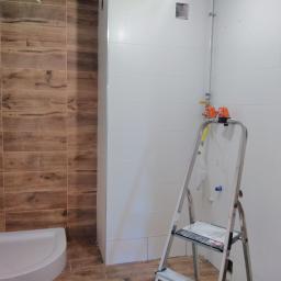 Jakub Kozyra Usługi Remontowe i Wykończeniowe - Wnętrze łazienki w trakcie remontu: ułożone płytki imitujące drewno na ścianie kabiny prysznicowej i podłodze, białe płytki na pozostałych ścianach, drabina aluminiowa z gazetą, widoczne rury...