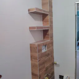 Wykończona łazienka z drewnopodobną zabudową, wnękami pełniącymi funkcję półek i białymi płytkami na ścianach, widoczne elementy instalacji hydraulicznej i elektrycznej.