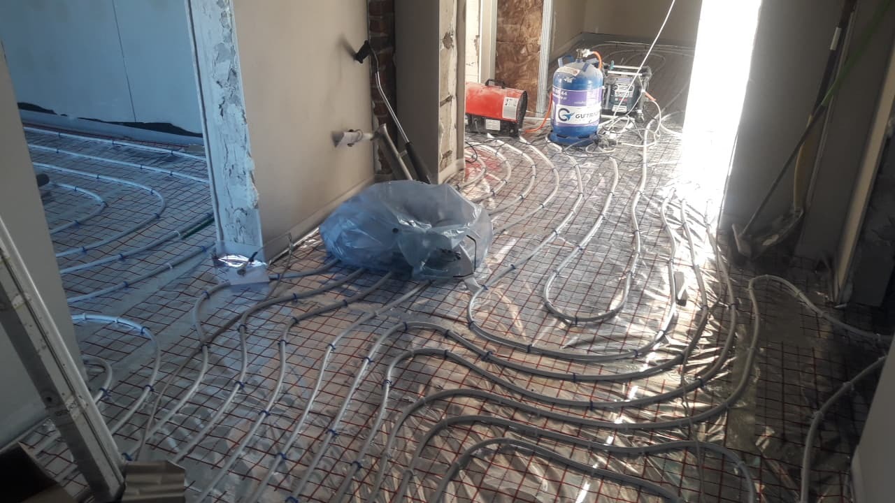 Instalacja ogrzewania podłogowego: białe rurki ułożone na folii z siatką, widoczny butla z gazem, nagrzewnica i worek foliowy.