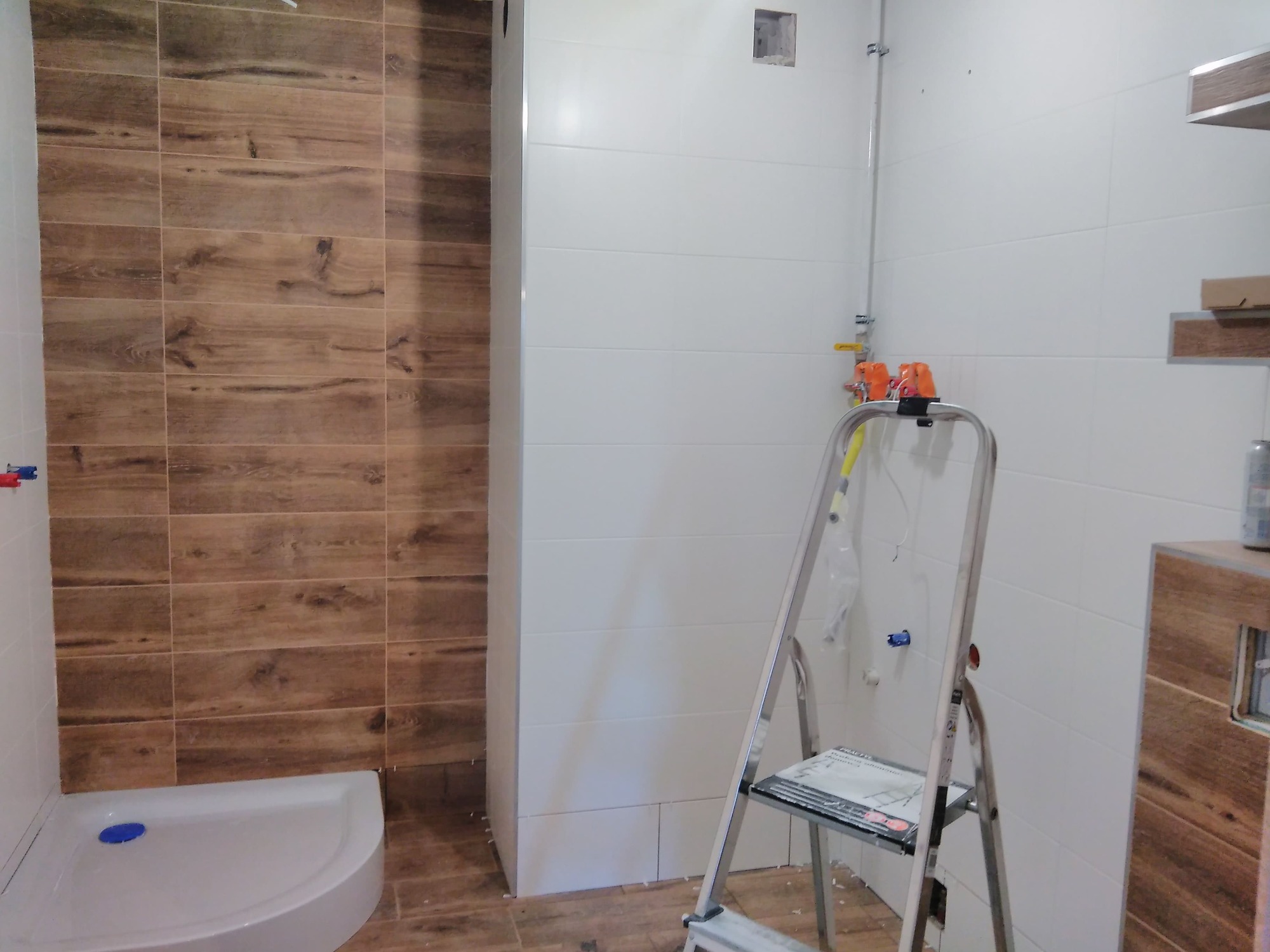 Wnętrze łazienki w trakcie remontu: ułożone płytki imitujące drewno na ścianie kabiny prysznicowej i podłodze, białe płytki na pozostałych ścianach, drabina aluminiowa z gazetą, widoczne rury...