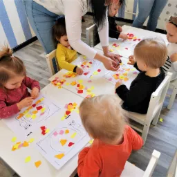 Grupa dzieci przy białych stolikach, wyklejających kolorowymi papierami obrazek ślimaka; nauczycielka pomaga jednemu z dzieci.