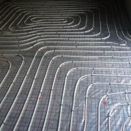 Sypex - Ułożone białe rury ogrzewania podłogowego na folii z czerwoną siatką, przymocowane czarnymi klipsami. Widok z góry na betonową podłogę, instalacja przed wylaniem posadzki.