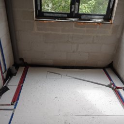 Sypex - Przygotowanie do ogrzewania podłogowego w surowej łazience: izolacja styropianowa, rury PEX czerwone i niebieskie prowadzone wzdłuż ścian, okno w tle.