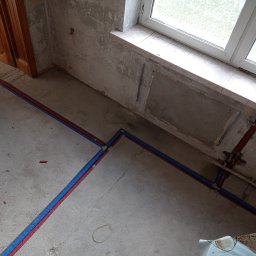 Sypex - Instalacja ogrzewania podłogowego z rur PEX w kolorach niebieskim i czerwonym, ułożonych na surowym betonie w remontowanym pomieszczeniu z oknem. Widoczne narzędzia i surowe ściany.