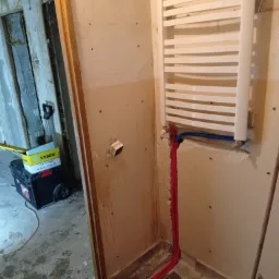 Biały grzejnik drabinkowy zamontowany na ścianie łazienki w trakcie remontu, widoczne rury doprowadzające wodę i narzędzia w tle.