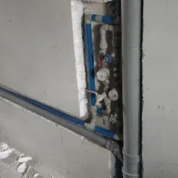 Wnęka w surowej ścianie z widocznymi, częściowo zamontowanymi rurami wodnymi, liczniki wody i pion kanalizacyjny. Na pierwszym planie drabina aluminiowa, wiadro i resztki materiałów budowlanych.