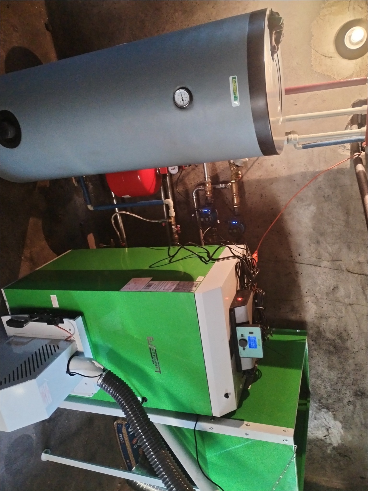 Zielony piec CO z szarym zasobnikiem na wodę, widoczne rury i zawory. Instalacja grzewcza w kotłowni. Układ z manometrem i pompami obiegowymi.