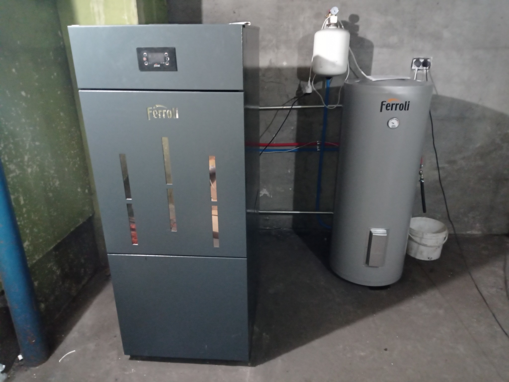 Szary piec CO marki Ferroli z bojlerem i osprzętem na tle szarej ściany. Widoczne rury instalacyjne i białe wiaderko. Ujęcie z bliska.