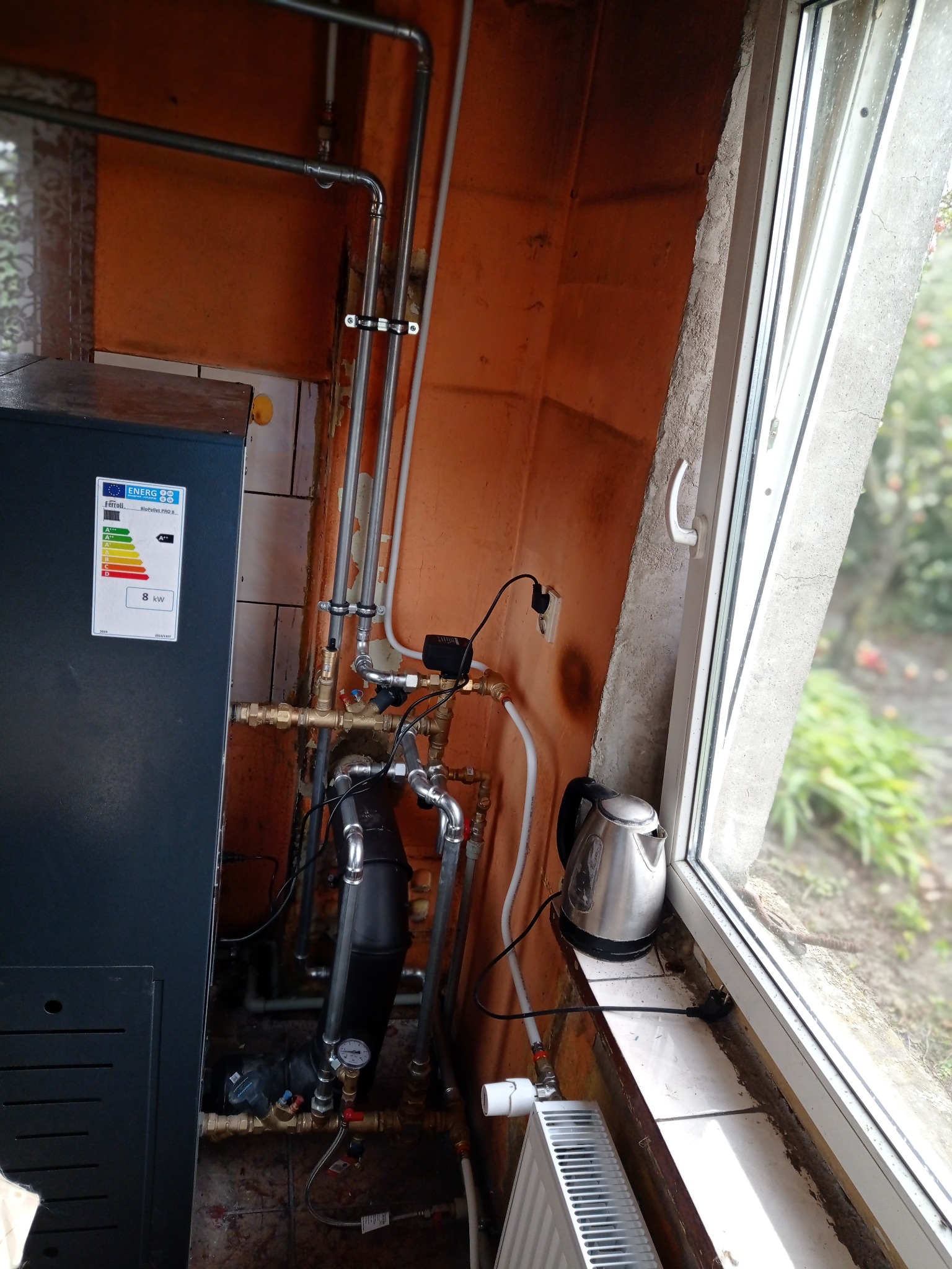 Kotłownia z piecem na pellet Ferroli BioPellet PRO 8 kW, rurami instalacji, grzejnikiem i czajnikiem elektrycznym przy oknie. Pomieszczenie z pomarańczowymi ścianami.