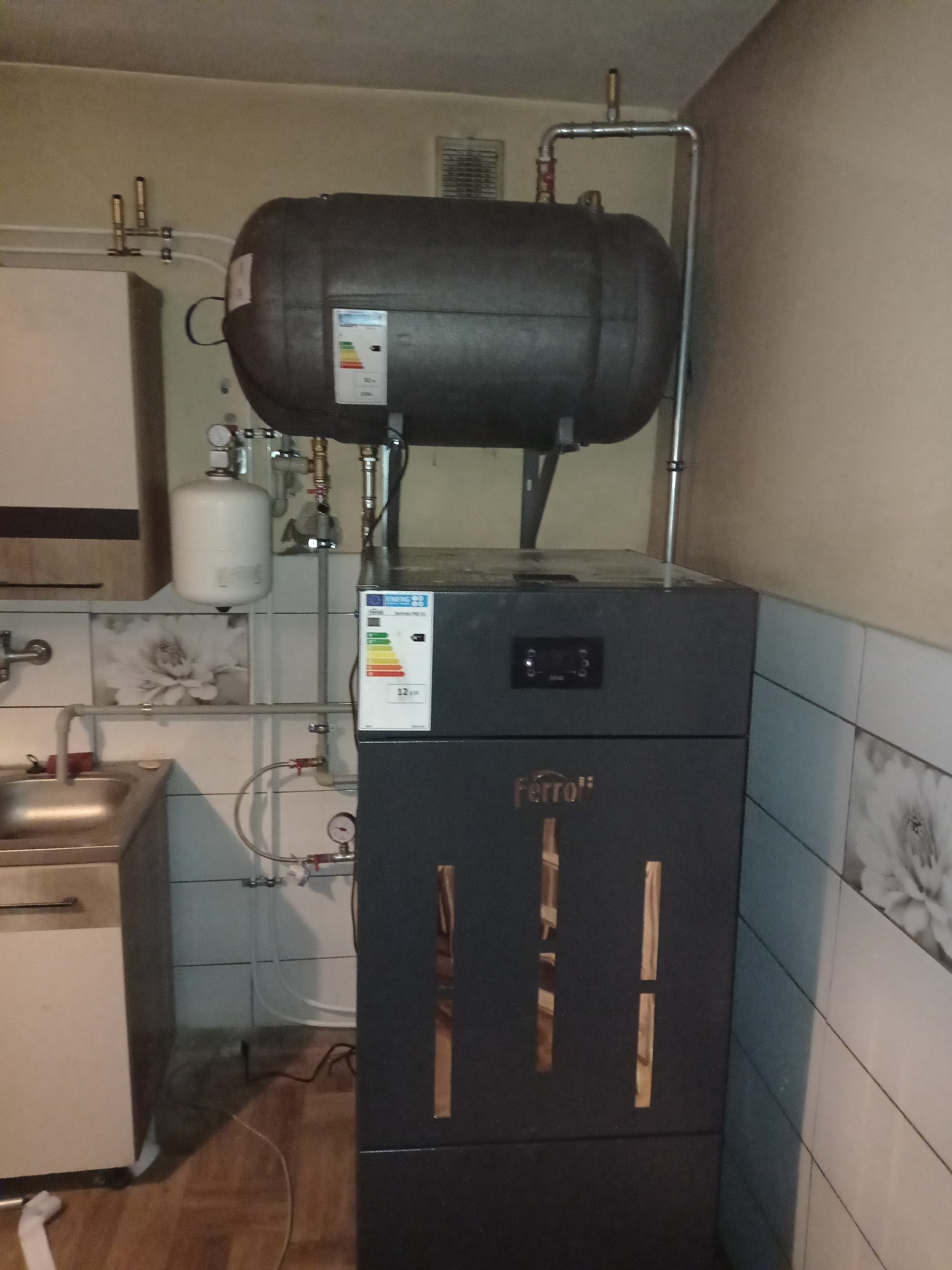 Zamontowany, nowoczesny piec CO marki Ferroli z zasobnikiem, widoczne rury instalacji grzewczej, manometry i etykieta energetyczna na tle ściany z płytek.