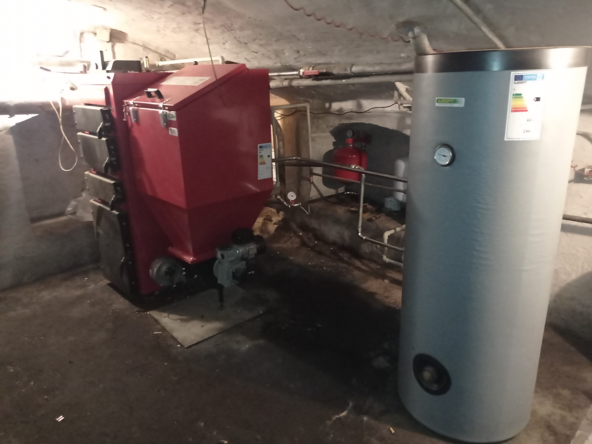 Czerwony piec CO z szarym zasobnikiem wody w piwnicy z rurami. Widoczne etykiety energetyczne i manometr. Instalacja grzewcza w surowym pomieszczeniu.