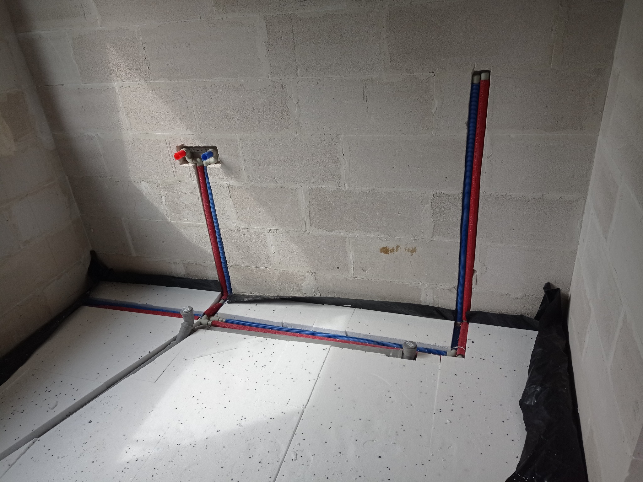 Instalacja ogrzewania podłogowego z rurami PEX w kolorze niebieskim i czerwonym, ułożonymi na białym styropianie, przy ścianie z betonu komórkowego.