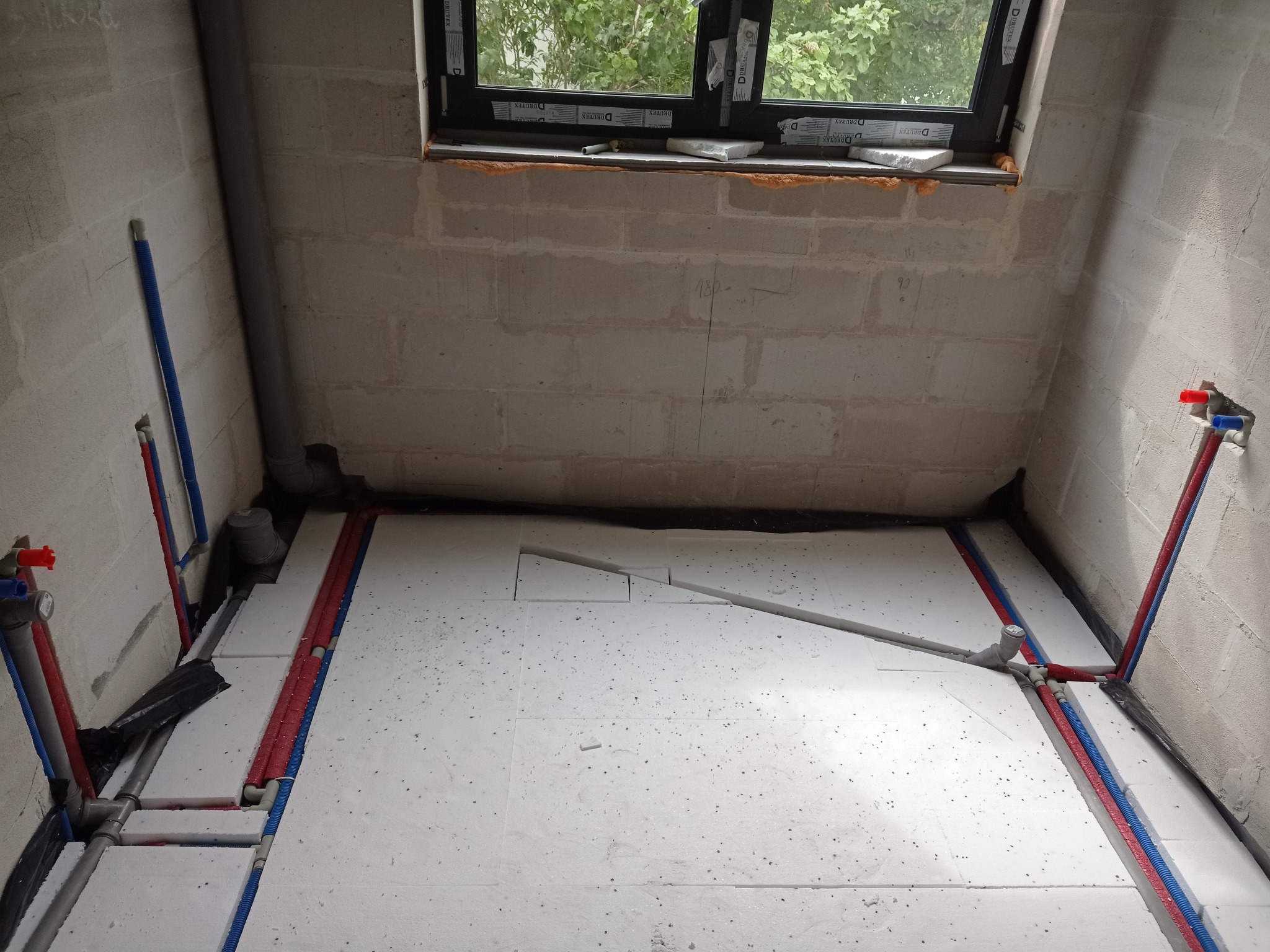 Przygotowanie do ogrzewania podłogowego w surowej łazience: izolacja styropianowa, rury PEX czerwone i niebieskie prowadzone wzdłuż ścian, okno w tle.