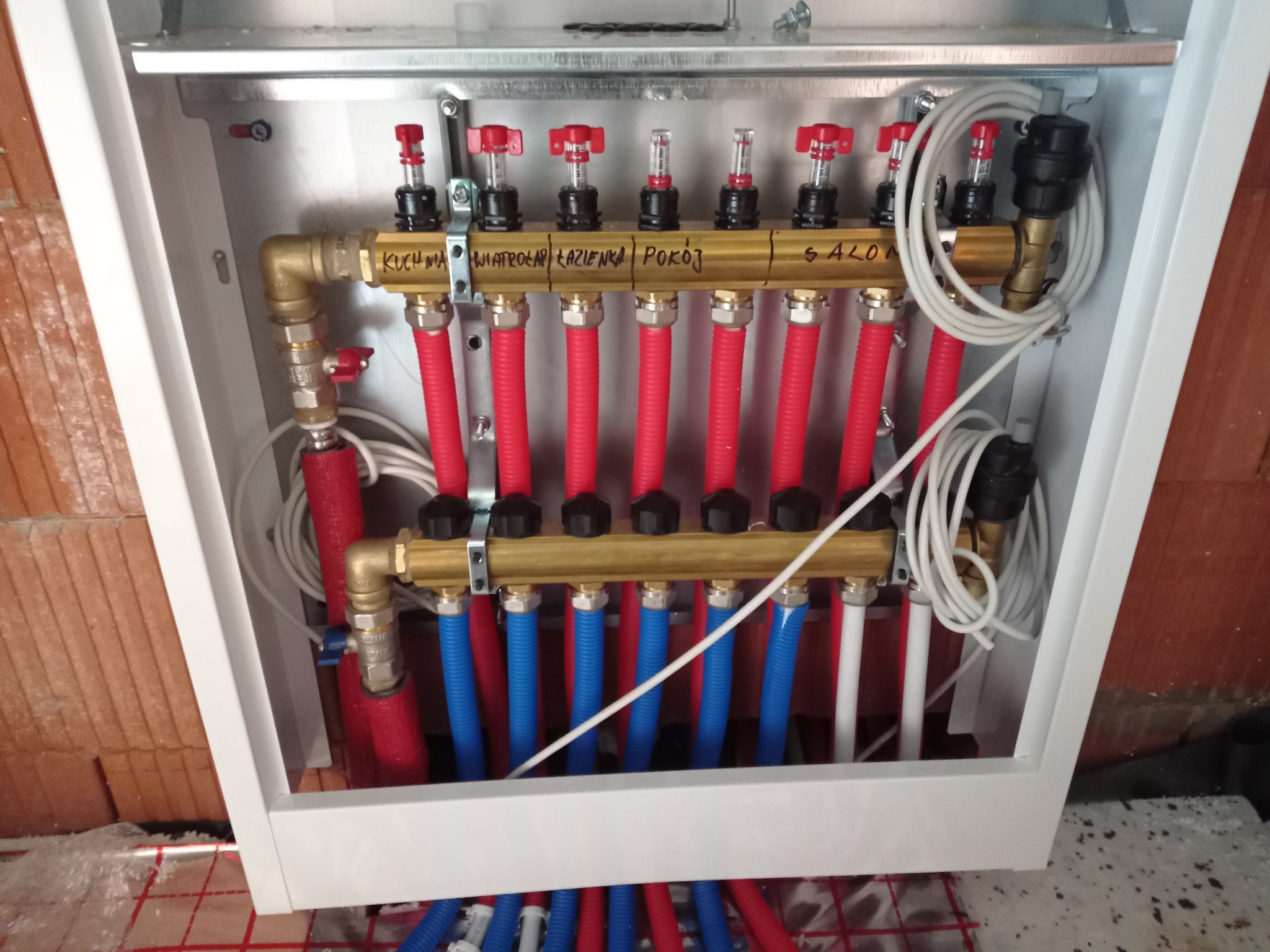 Rozdzielacz ogrzewania podłogowego w białej szafce, z rurami oznaczonymi nazwami pomieszczeń (kuchnia, wiatrołap, łazienka, pokój, salon).
