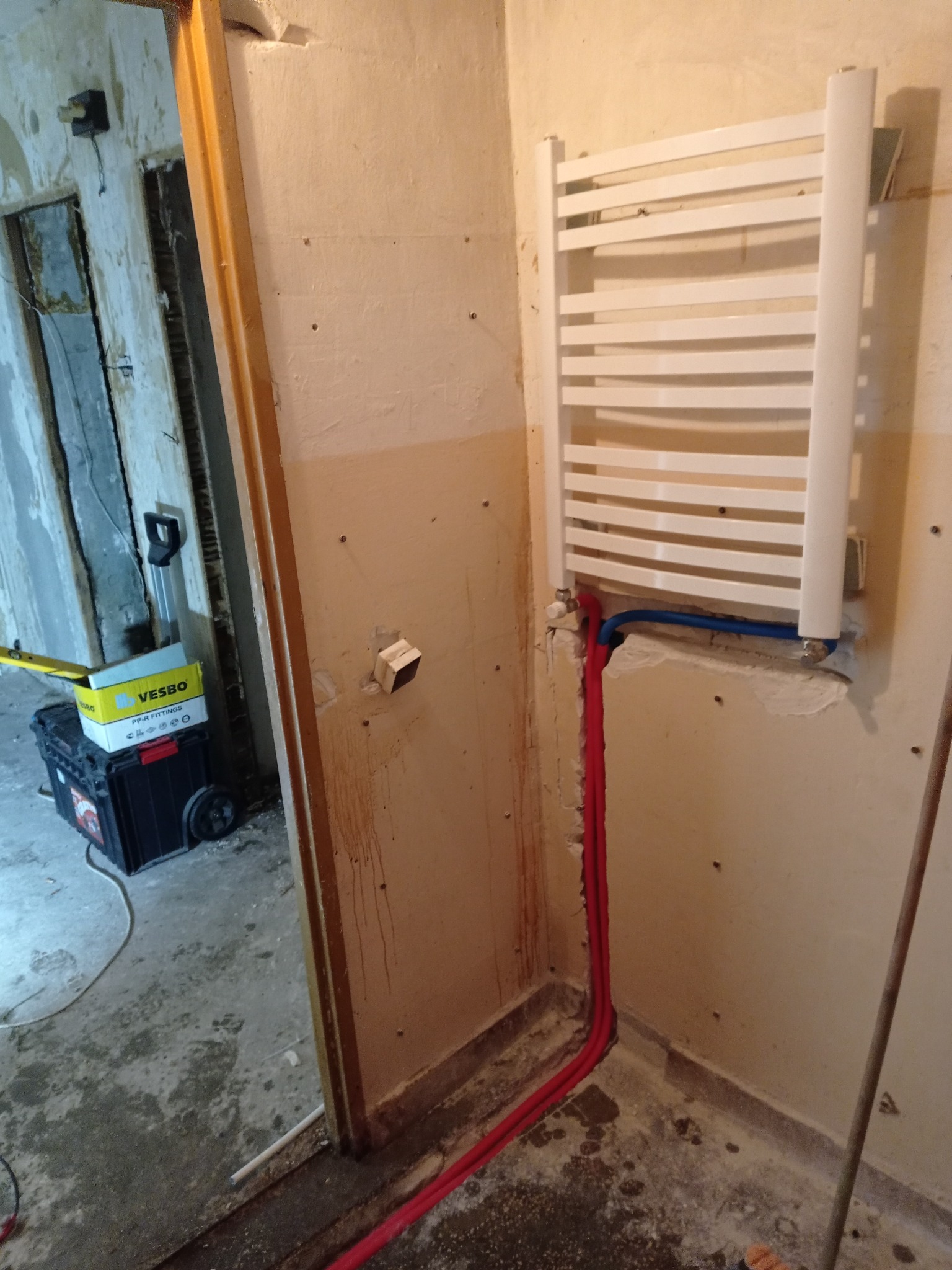 Biały grzejnik drabinkowy zamontowany na ścianie łazienki w trakcie remontu, widoczne rury doprowadzające wodę i narzędzia w tle.