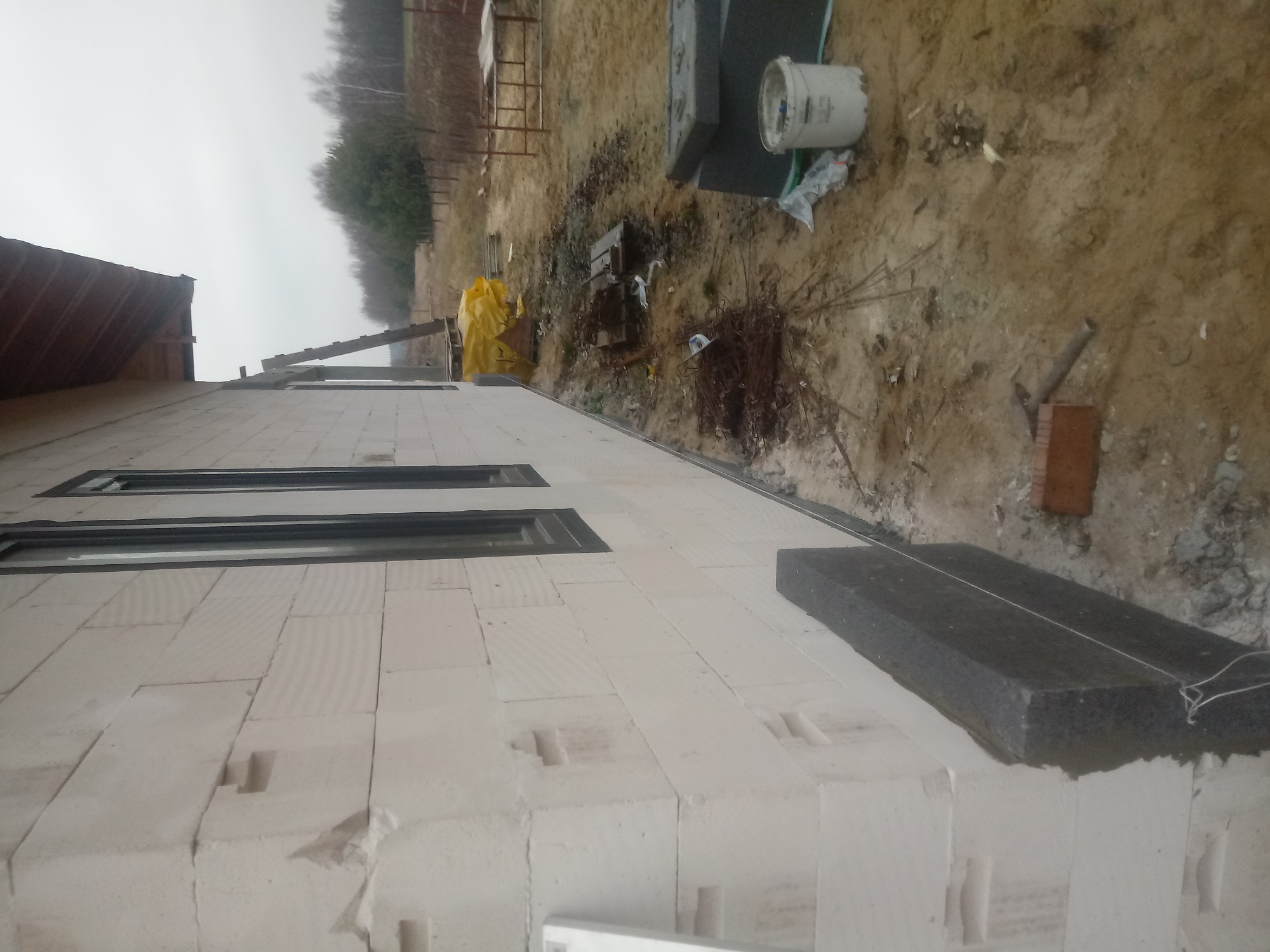 Ściana domu w trakcie budowy z białych bloczków, widoczne okna i fragment ocieplenia, na ziemi wiadro i narzędzia.