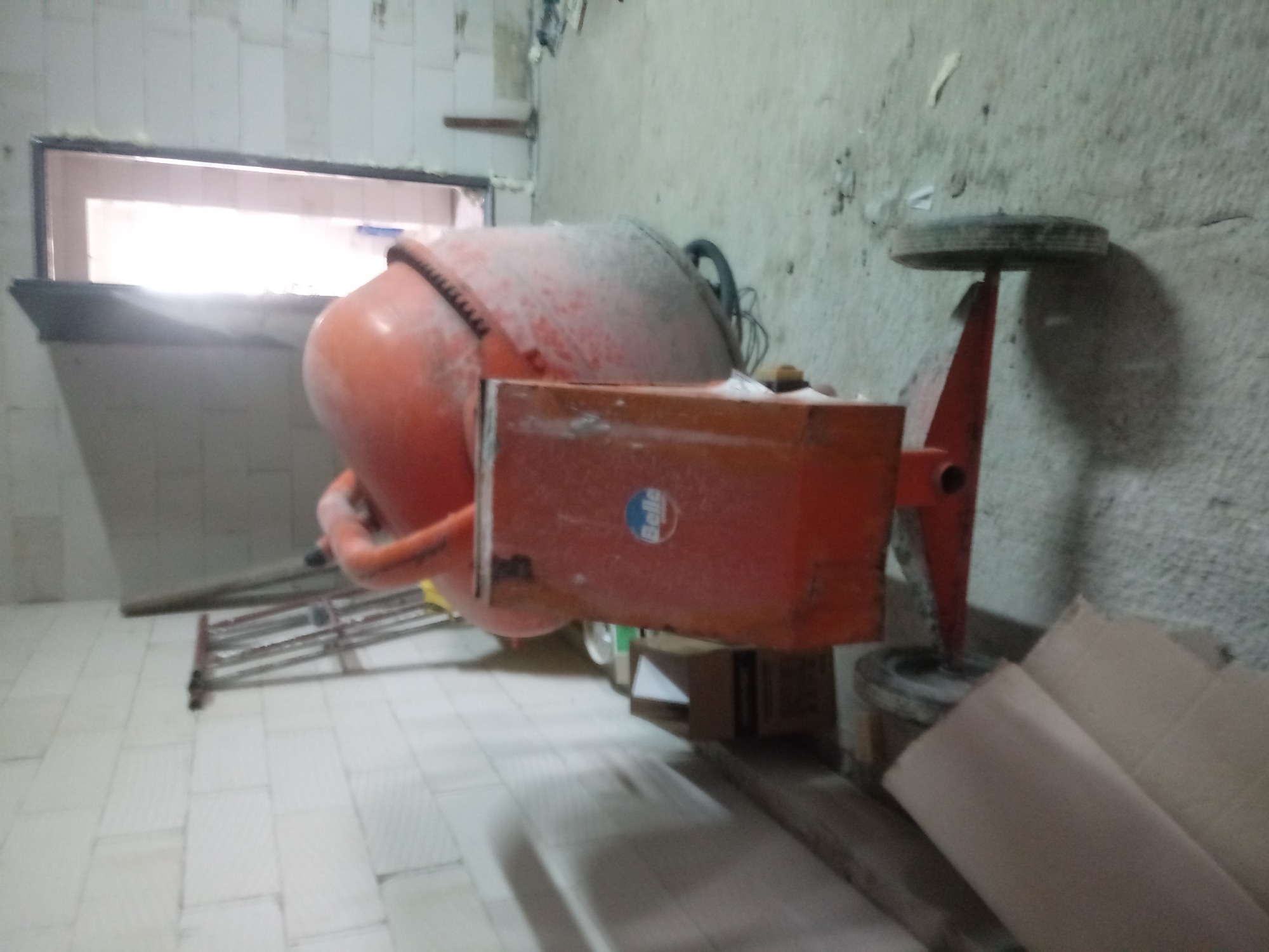 Pomarańczowa betoniarka marki Belle, lekko zabrudzona, stoi na podłodze z płytek w pomieszczeniu z oknem i drabiną w tle.