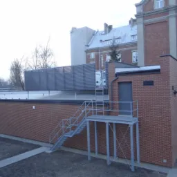 Nowo wybudowany budynek gospodarczy z cegły z metalowymi schodami zewnętrznymi i platformą, widoczny na tle starszego budynku z cegły.
