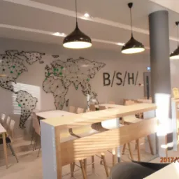 Jasne biuro z mapą świata na ścianie, wiszącymi lampami w kształcie dzwonów i minimalistycznymi meblami w odcieniach bieli i drewna.