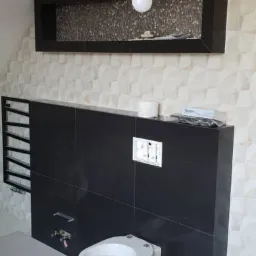 Wnętrze łazienki w trakcie remontu: czarne płytki ścienne, podwieszana toaleta, dekoracyjna ściana z trójwymiarowymi płytkami, wisząca półka z mozaiką i żarówka na kablu.