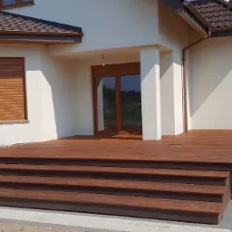 Nowoczesny dom z tarasem z drewna egzotycznego i schodami, brązowe rolety zewnętrzne, jasna elewacja i kostka brukowa w szarościach.