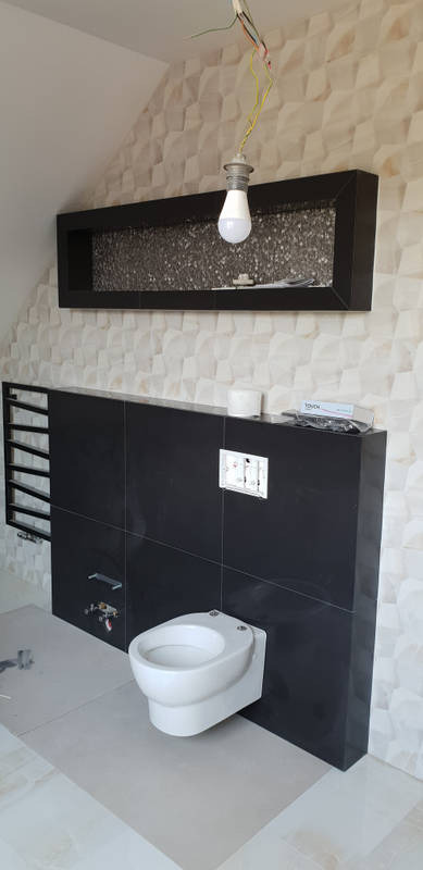 Wnętrze łazienki w trakcie remontu: czarne płytki ścienne, podwieszana toaleta, dekoracyjna ściana z trójwymiarowymi płytkami, wisząca półka z mozaiką i żarówka na kablu.