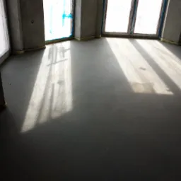 Szara wylewka podłogowa w surowym wnętrzu, promienie słoneczne wpadają przez okna, widoczne szare ściany i żółte zabezpieczenia przy oknach.