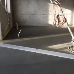 Świeżo wylana, szara posadzka w surowym wnętrzu, wyrównywana łatą aluminiową i obrabiana mechaniczną zacieraczką do betonu.