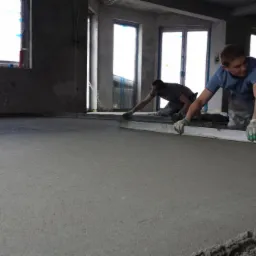 Dwóch mężczyzn w rękawicach wyrównuje świeżo wylaną posadzkę jastrychową przy użyciu długiej łaty w surowym wnętrzu z oknami.