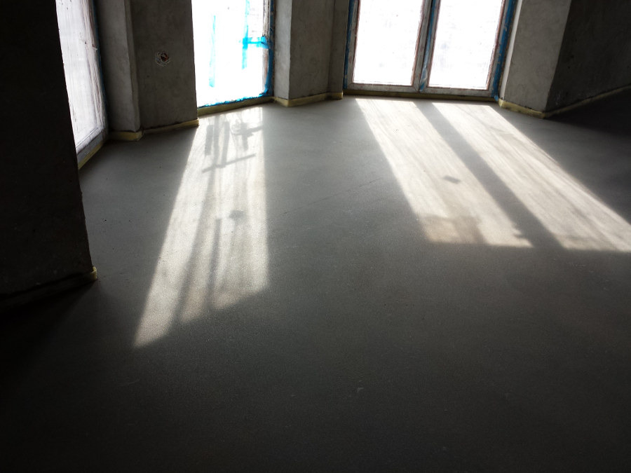 Szara wylewka podłogowa w surowym wnętrzu, promienie słoneczne wpadają przez okna, widoczne szare ściany i żółte zabezpieczenia przy oknach.