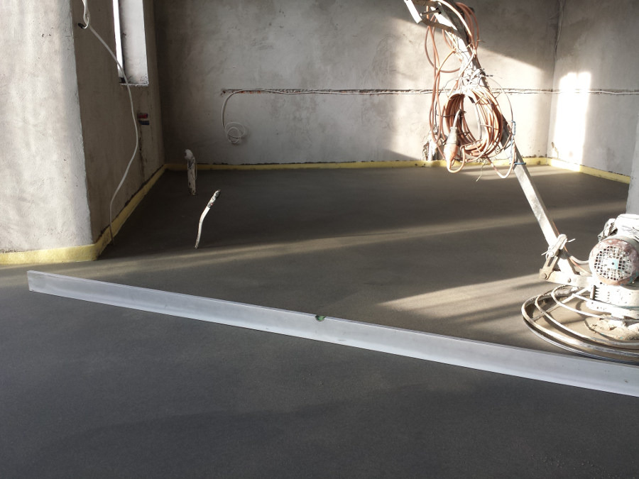 Świeżo wylana, szara posadzka w surowym wnętrzu, wyrównywana łatą aluminiową i obrabiana mechaniczną zacieraczką do betonu.