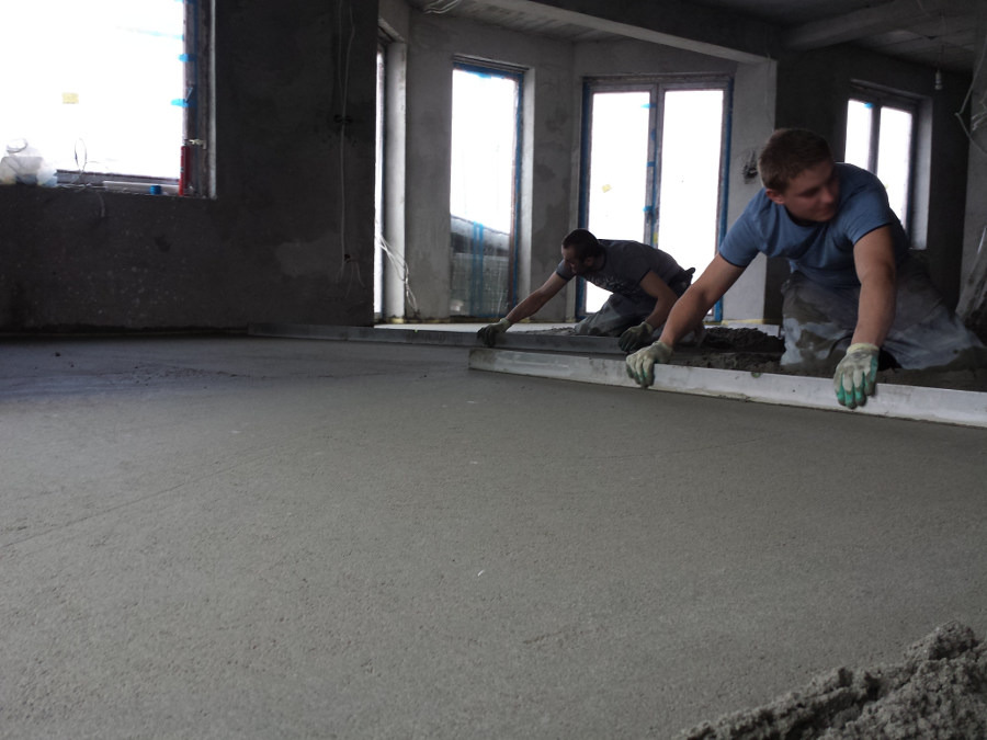 Dwóch mężczyzn w rękawicach wyrównuje świeżo wylaną posadzkę jastrychową przy użyciu długiej łaty w surowym wnętrzu z oknami.