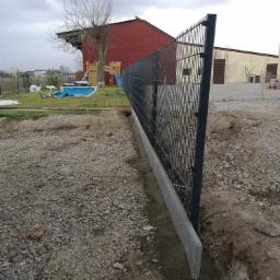 Montaż ogrodzenia panelowego na betonowej podmurówce, widoczne wykopane rowy i gruz, w tle budynki gospodarcze i plac zabaw.