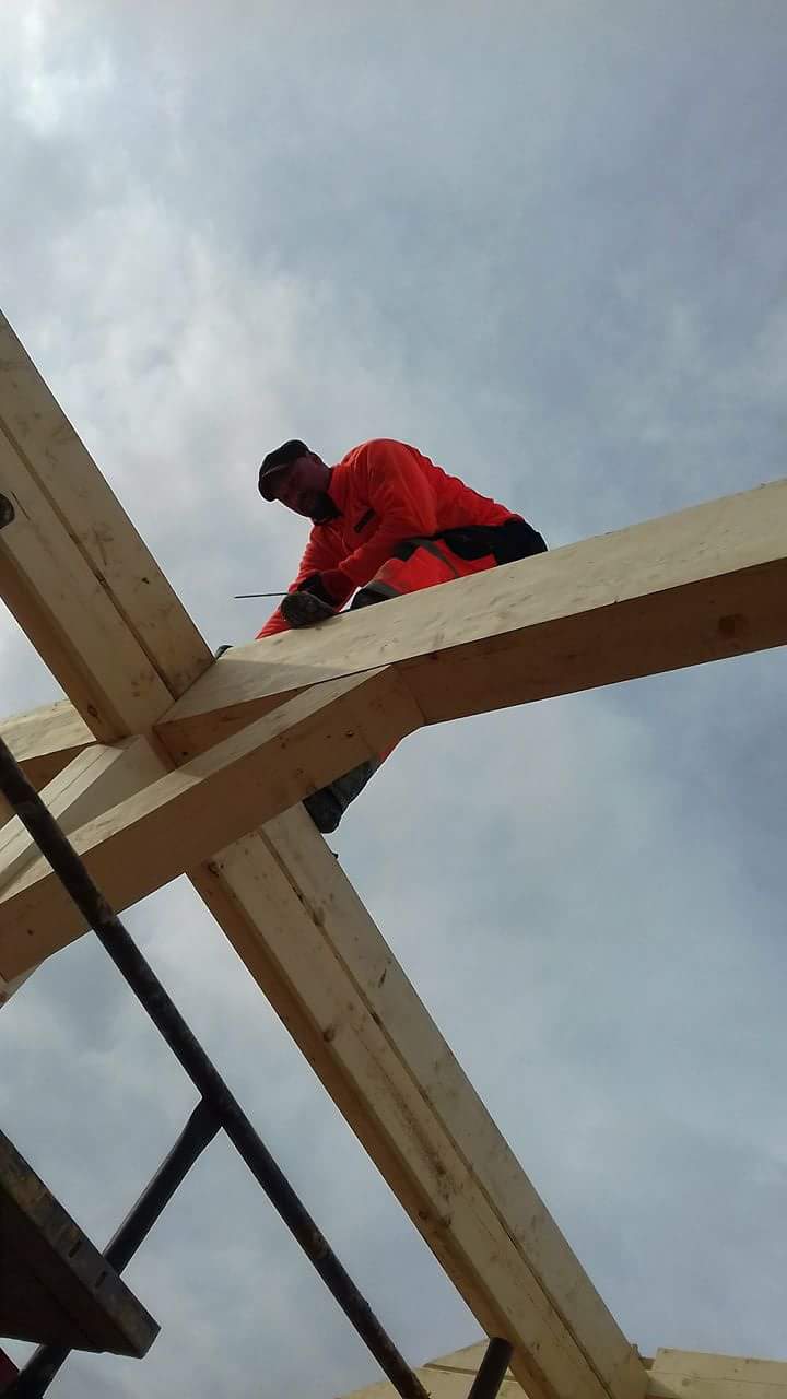 Monter w pomarańczowym kombinezonie łączy elementy drewnianej konstrukcji domu szkieletowego na tle pochmurnego nieba.