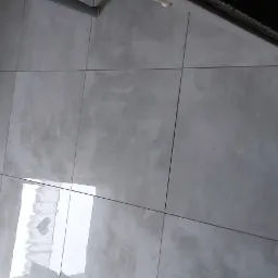 Szare, matowe płytki ceramiczne na ścianie i podłodze w łazience, widoczny fragment listwy przypodłogowej i rury.