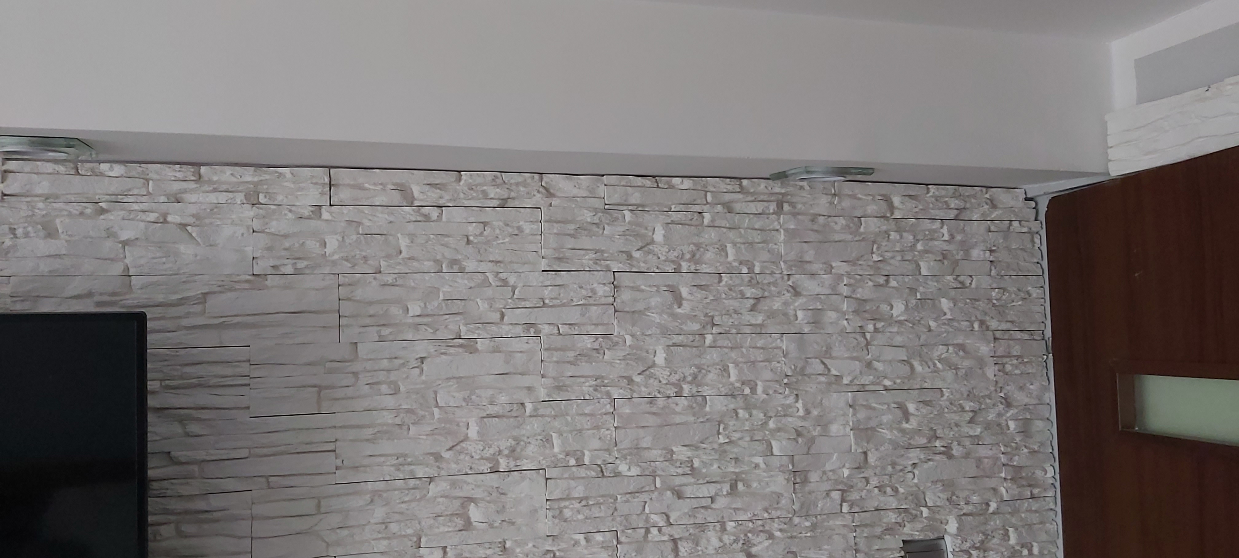 Ściana wyłożona dekoracyjną, białą cegłą z widocznymi punktowymi lampami sufitowymi i fragmentem ciemnobrązowych drzwi z mlecznym szkłem.