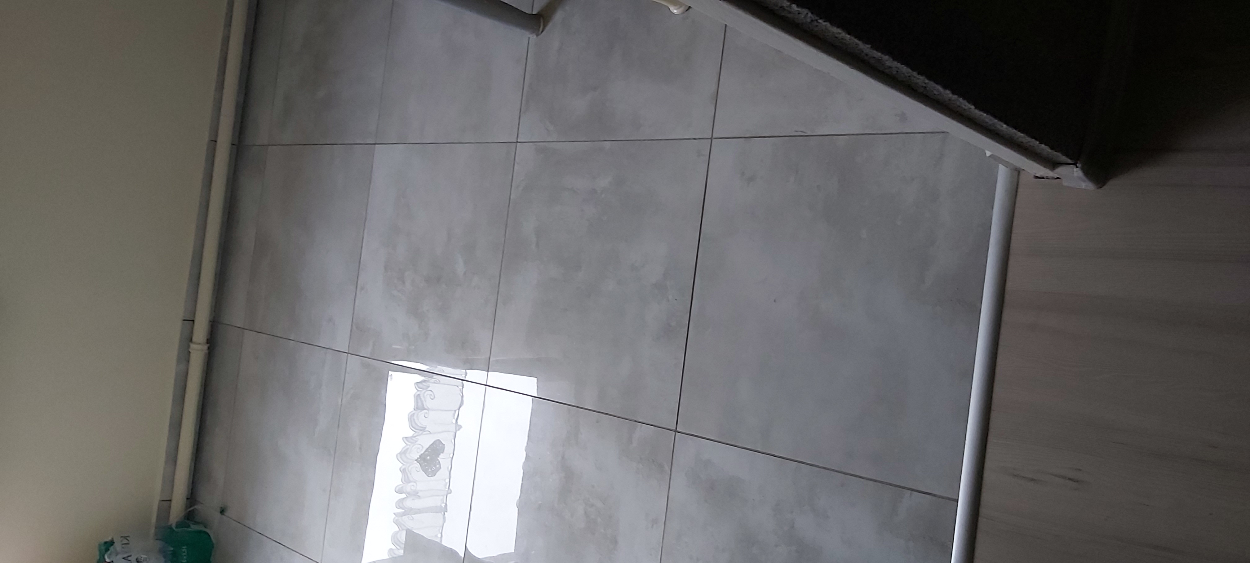 Szare, matowe płytki ceramiczne na ścianie i podłodze w łazience, widoczny fragment listwy przypodłogowej i rury.