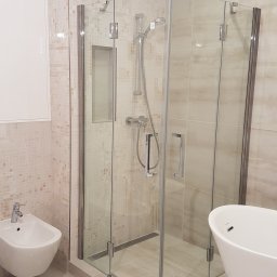 Zuko Sp.zo.o - Nowoczesna kabina prysznicowa z transparentnego szkła, chromowanymi zawiasami i odpływem liniowym, obok białe bidet i wanna wolnostojąca, całość w łazience wykończonej beżowymi płytkami w dwóch...