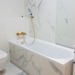 Zuko Sp.zo.o - Biała wanna obudowana płytkami imitującymi marmur, złota bateria i deszczownica, ściany wyłożone białymi płytkami z szarymi żyłami, widoczny fragment toalety