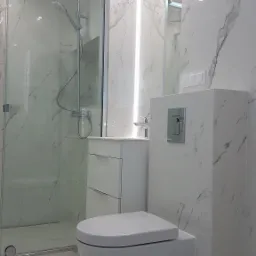 Nowoczesna łazienka wykończona białym marmurem, z podwieszaną toaletą, kabiną prysznicową ze szklanymi drzwiami i szafką z umywalką, oświetlona punktowymi lampami sufitowymi i paskiem LED.