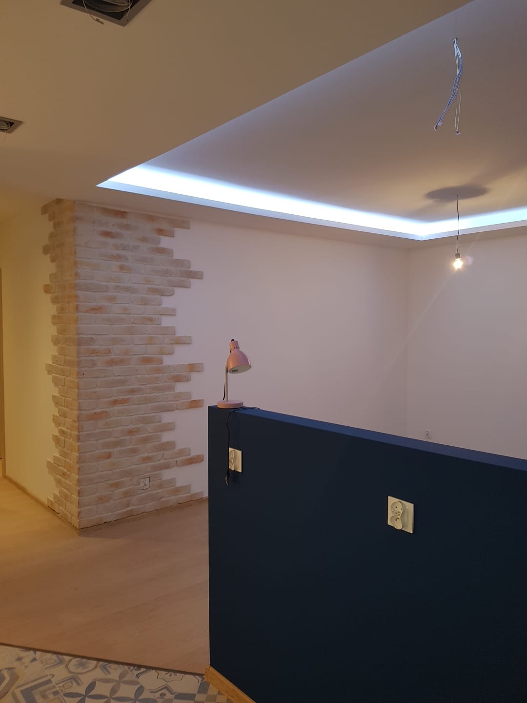 Wykończone wnętrze we Wrocławiu z dekoracyjną ścianą z cegły, listwą LED w suficie podwieszanym, granatową ścianką działową i widocznymi gniazdkami elektrycznymi.