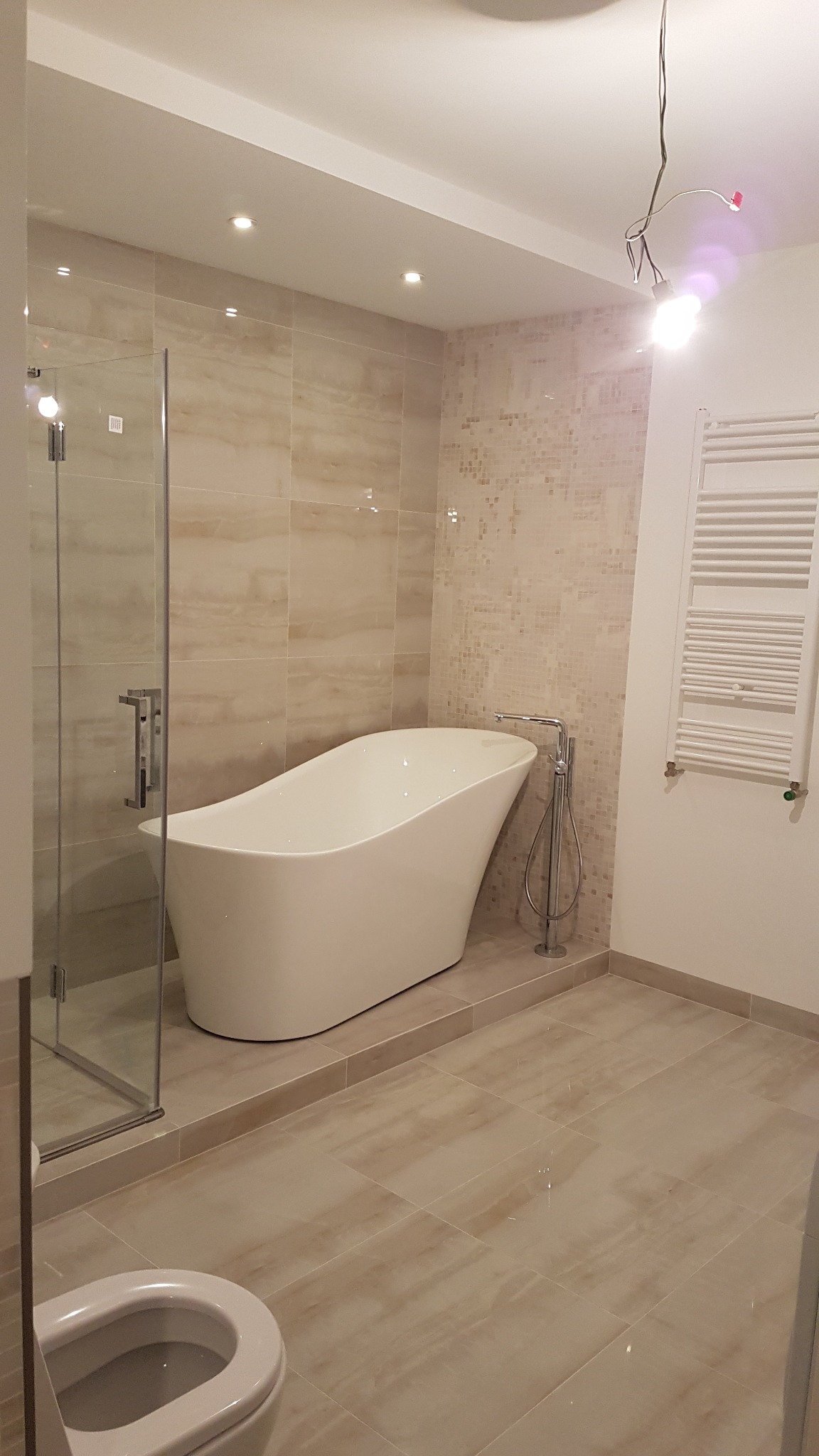 Nowoczesna łazienka z wolnostojącą wanną, kabiną prysznicową ze szklanymi drzwiami, grzejnikiem drabinkowym i beżowymi płytkami na ścianach i podłodze. Niedokończone oświetlenie sufitowe...